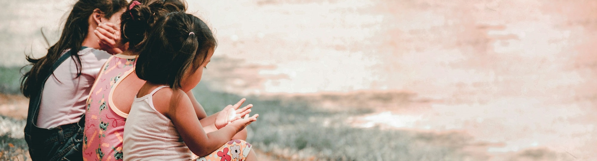 Three children sitting in the grass on the left side of the image. Photo by Charlein Gracia via UnSplash